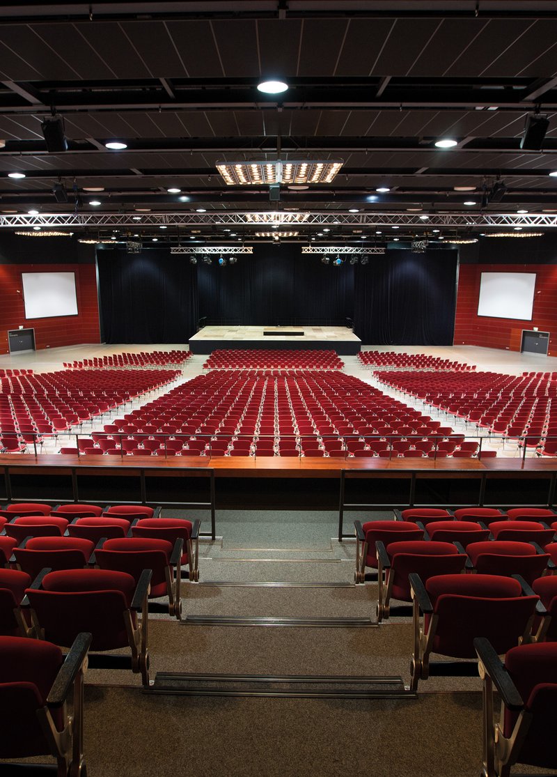 Blick die Tribüne hinunter in den Großen Saal des RuhrCongress Bochum. Rote Stuhlreihen säumen die Tribüne. Im Hintergrund steht eine Bühne vor einem schwarzen Vorhang und links und rechts davon hängen Leinwände.