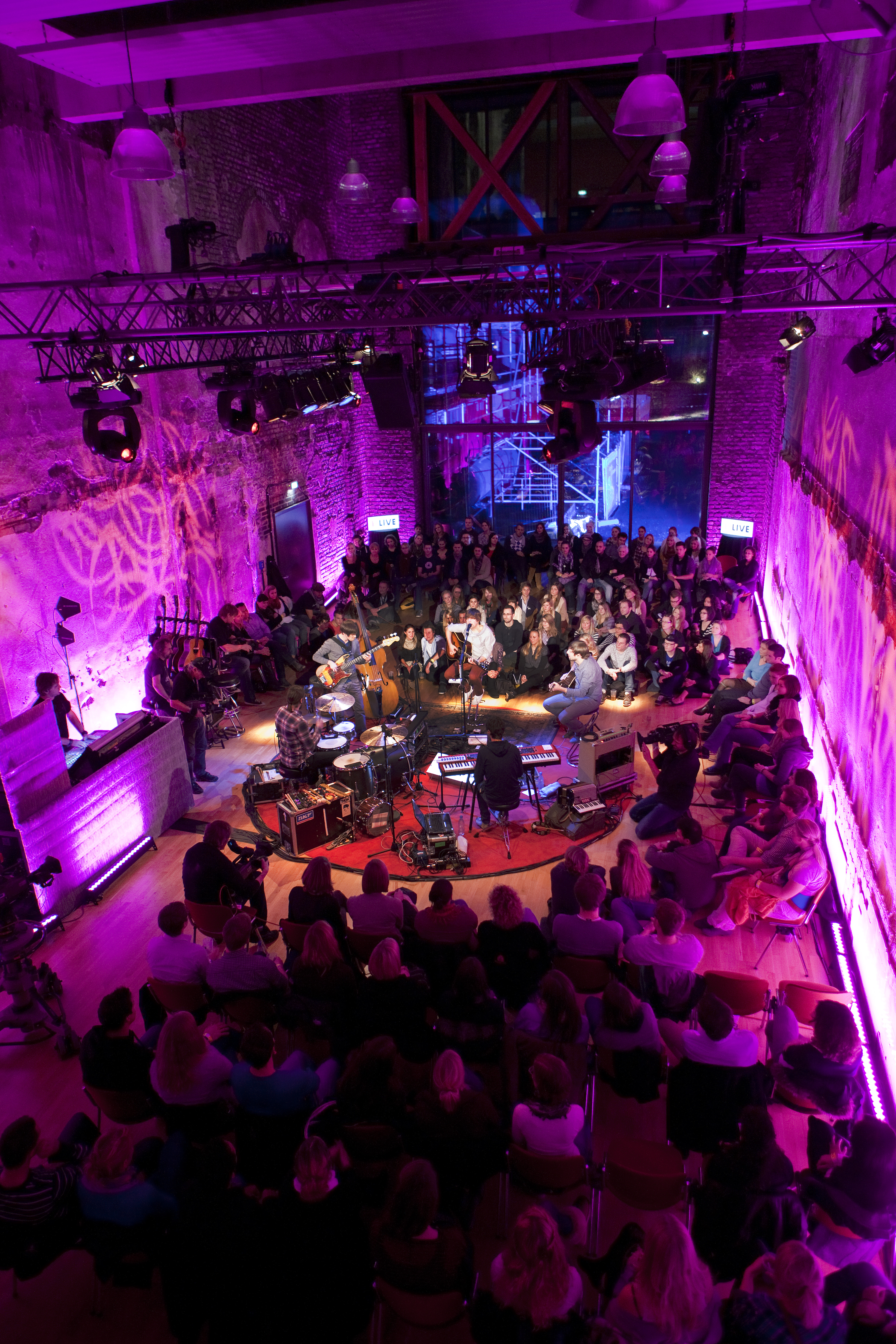 Besucher:innen einer Veranstaltung im Pumpenhaus an der Jahrhunderthalle Bochum sitzen um eine runde Bühne herum und lauschen einer 4-köpfigen Band. Der Raum ist pink beleuchtet.