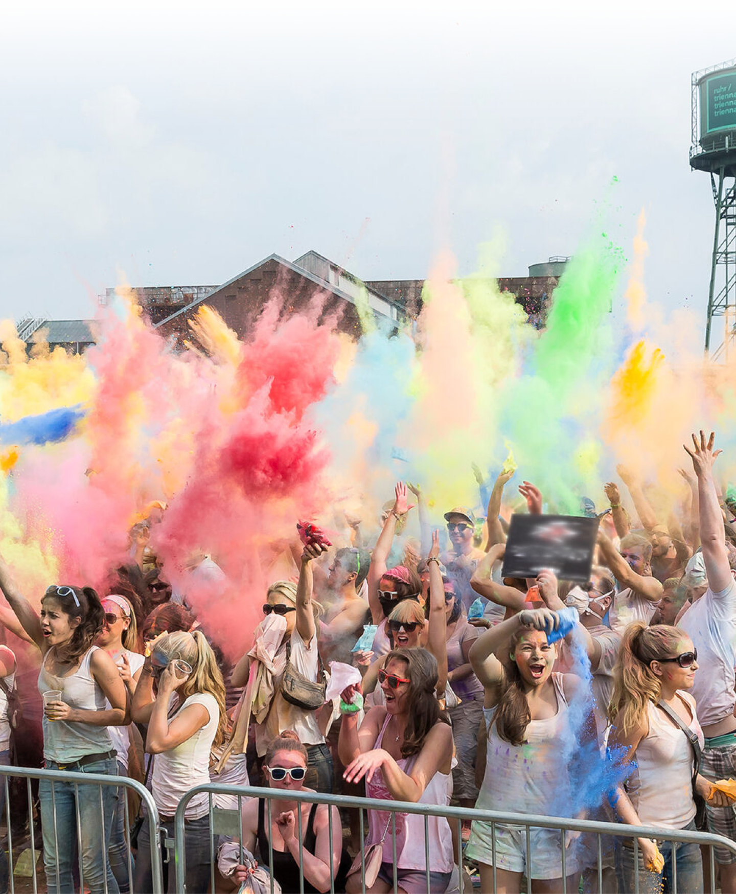 Menschen tanzen ausgelassen beim Holi-Festival auf der Nordwiese. Farbpulver wird in die Luft geworfen. Im Hintergrund ist die Jahrhunderthalle Bochum zu sehen.